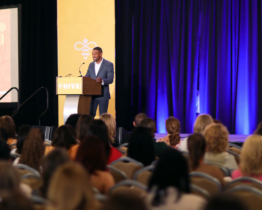 Jason Isaacs stands at the podium addressing an attentive audience at a HIVE 2023.