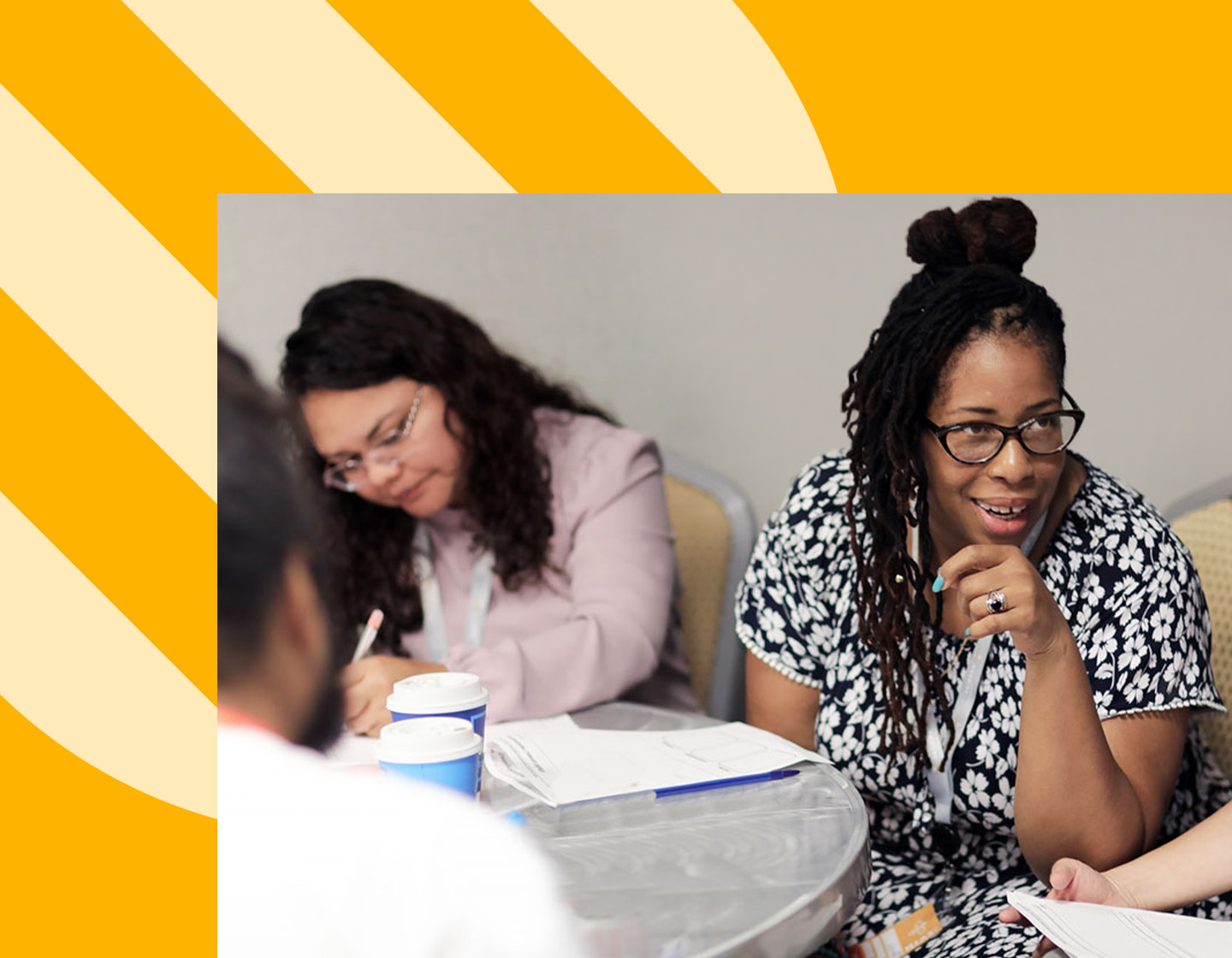 A focused, dark skin toned educator listens to another educator at a HIVE workshop where there are papers and coffee on a communal table.