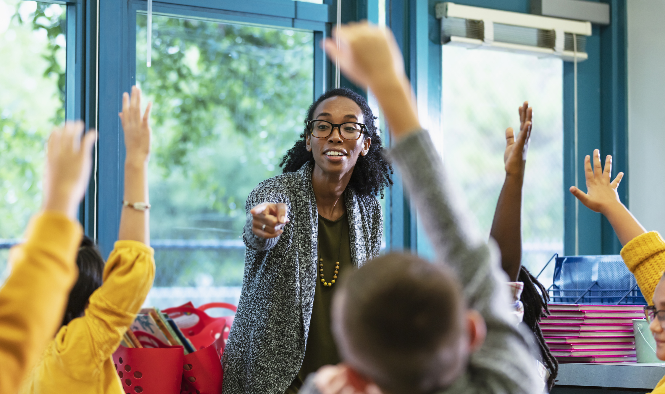 A multiracial group of elementary students in class raising their hands to answer a question. The focus is on their teacher, a dark skin toned woman in her 30s, who is standing in front, facing them, smiling and pointing to a student. Most of the students are 9 years old.