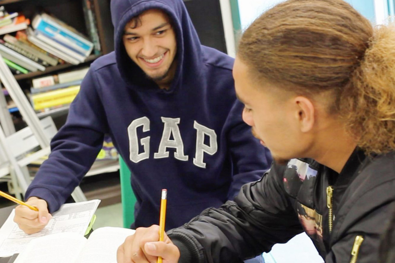 Two students engaged in a lighthearted study session, with one person wearing a hoodie smiling as they look at their companion who is focused on writing.