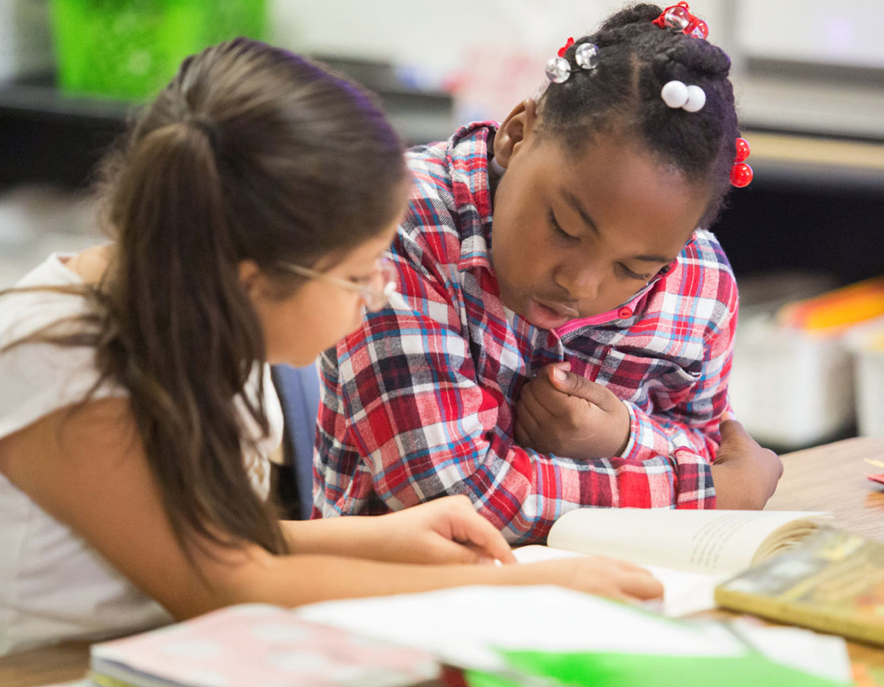 Two young students, one a medium skin toned student in pig tails, the other a dark skin toned student with a beaded hairstyle, are engrossed in reading a book together in a bright classroom setting.
