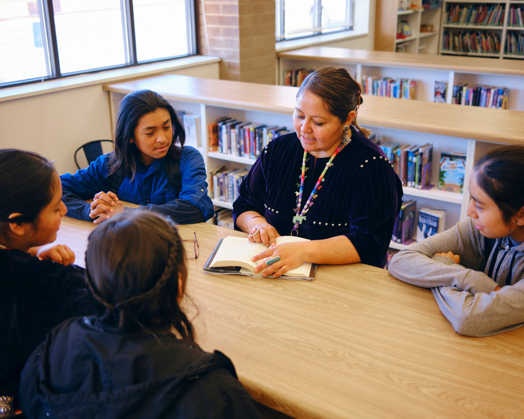 A teacher wearing a traditional indigenous garment and jewelry shares a story with her attentive students in a cozy library setting.