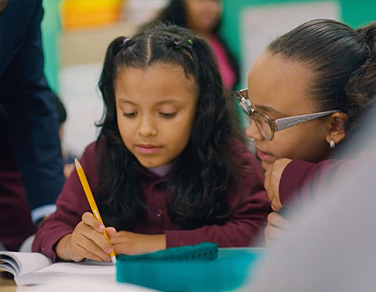 Two lighter dark skin toned students focusing intently on their schoolwork; one writing rigorously in their notebook while their companion observes meticulously.