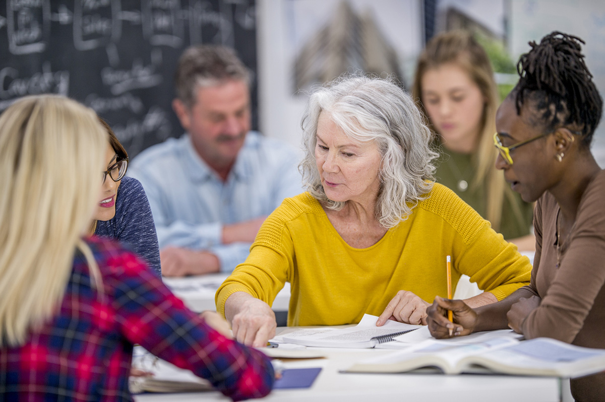 A diverse group of adult professionals of various ages engaged in a collaborative learning session, with an older woman leading the discussion while others listen attentively.