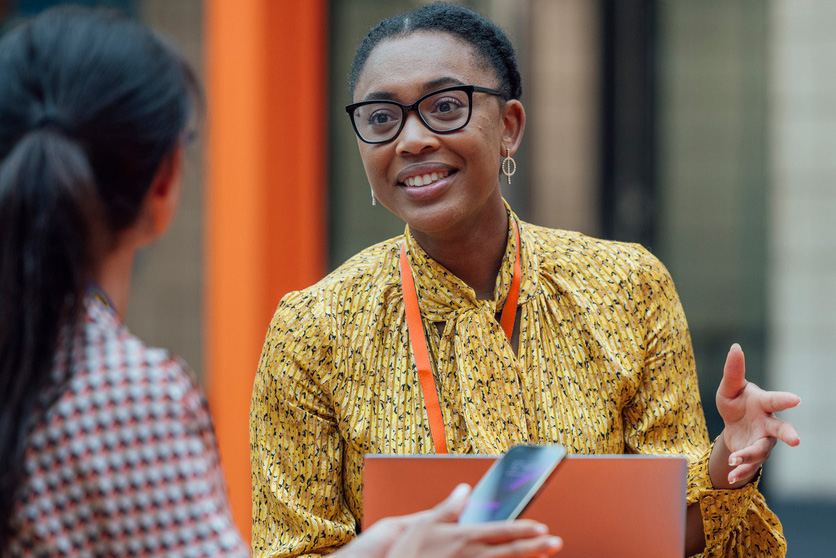 Two professionals engaging in a friendly and animated conversation at a networking event or conference, one is a dark skin toned person with an orange lanyard and bright yellow shirt.