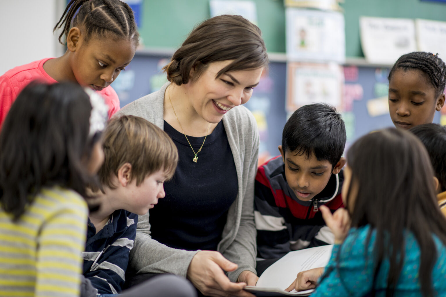 A multi-ethnic group of elementary school children wearing casual clothing are gathered around their teacher, who is light skinned and wearing a grey sweater, to listen during story time.